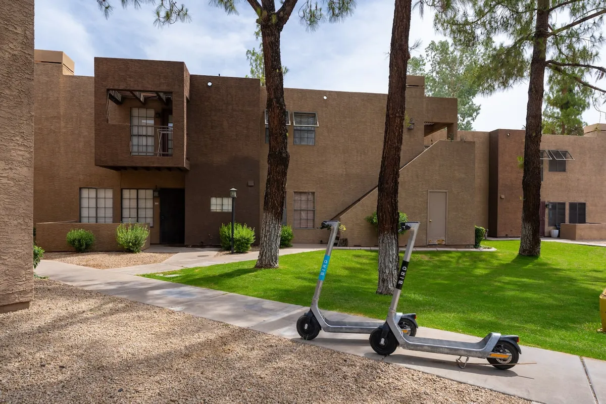 A casual outdoor setting with a scooter parked on a green lawn, shaded by trees and fronted by a building facade. The scene captures the essence of movement and neighborhood exploration, with a leisurely vibe.