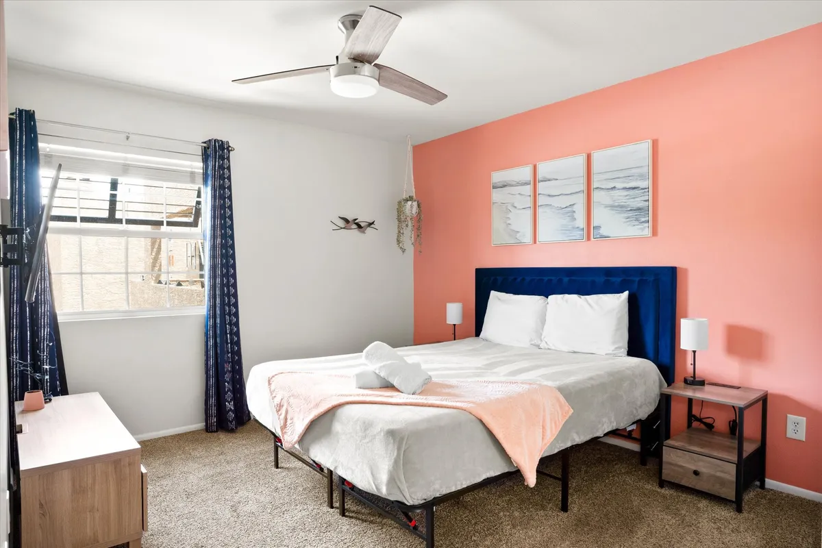 A tranquil bedroom featuring a bed with a blue headboard, a coral accent wall, framed artwork, and nightstands with lamps. The space is filled with natural light, creating an inviting ambiance for rest and relaxation.