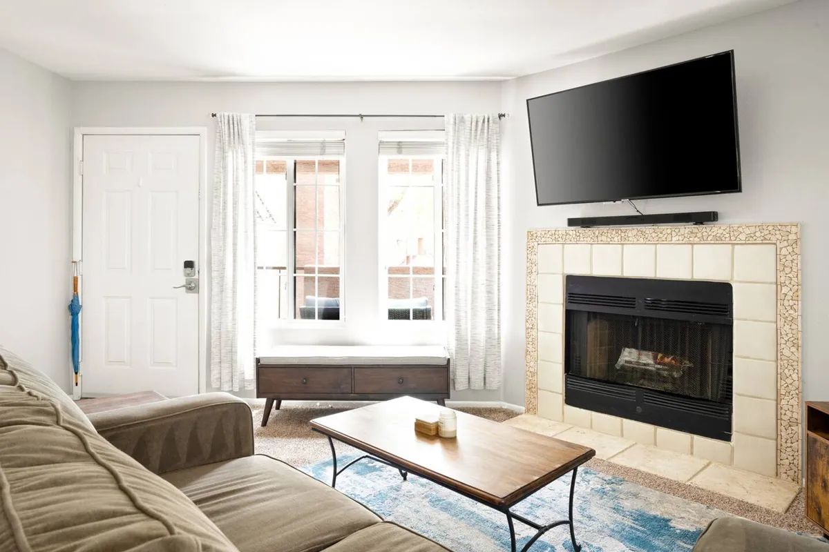 The image centers on a cozy living area with a plush sofa, fireplace, coffee table, and a large window seat. A television is mounted nearby, suggesting streaming-ready entertainment, and sunlight pours in through the windows, balanced by window coverings to ensure privacy.