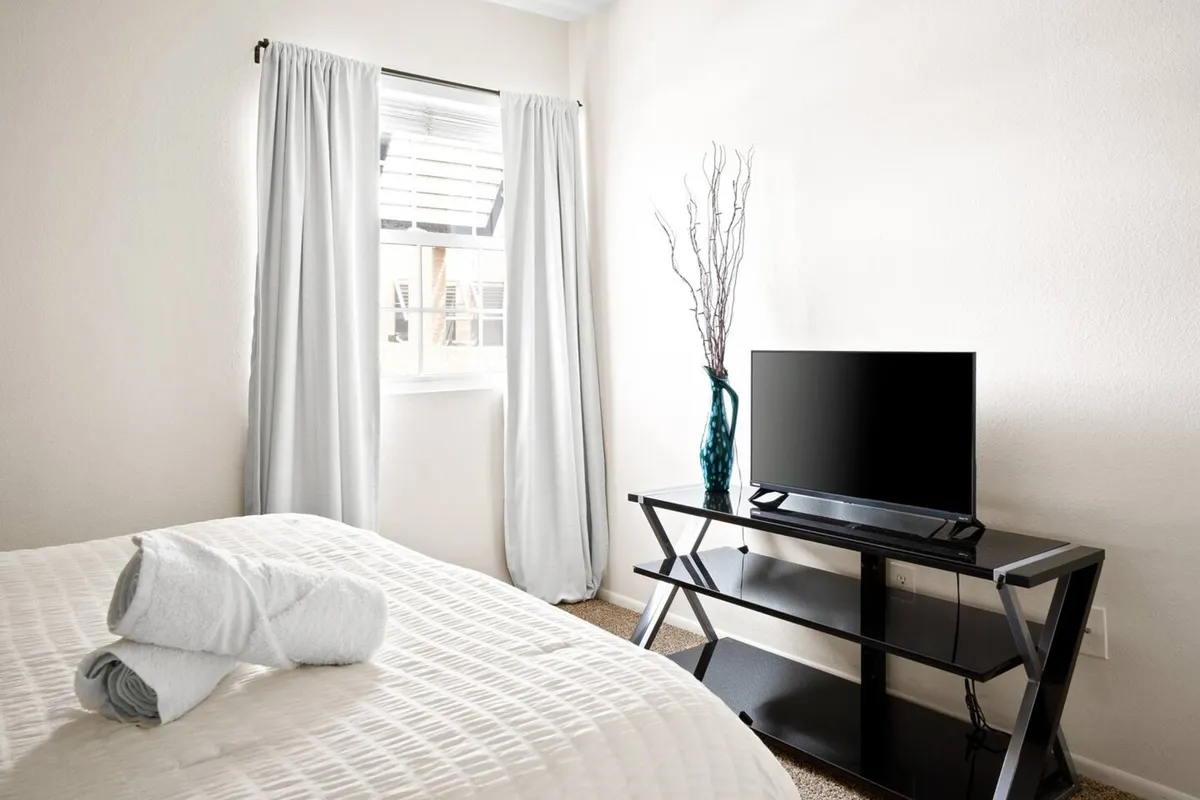 A peaceful bedroom scene with a bed adorned with rolled towels, a television on a black stand, a decorative branch in a vase, and soft curtains letting in natural light. The mood is quiet and restful.