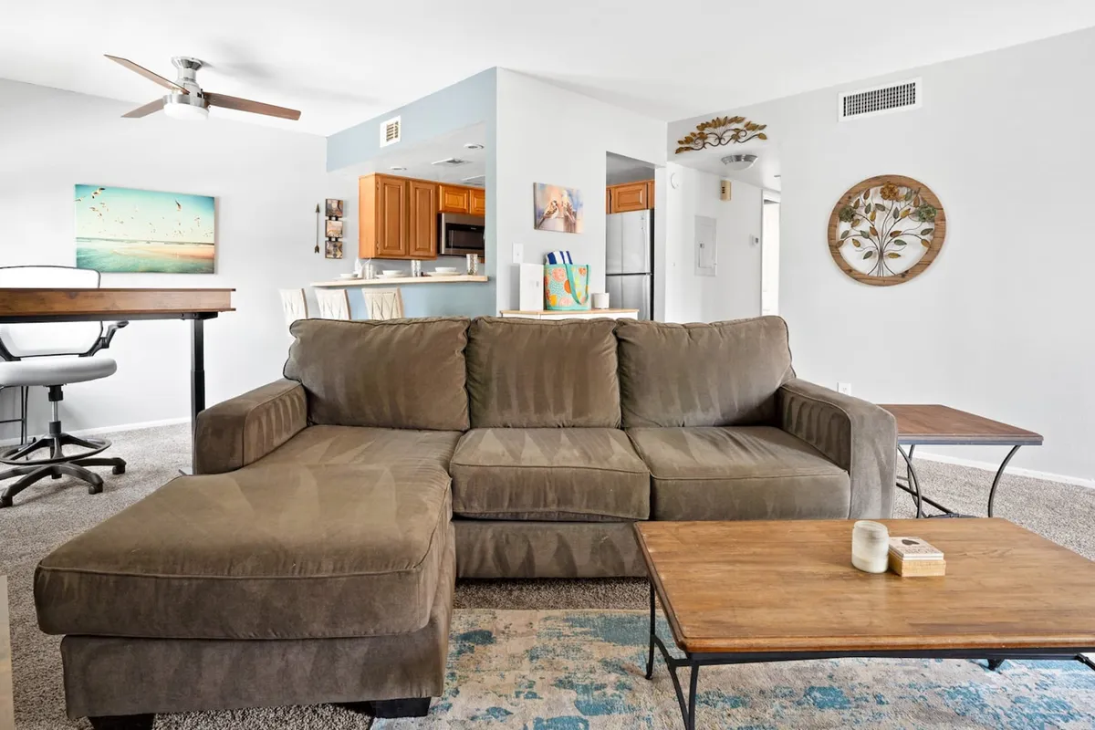 This image showcases a comfortable sectional sofa arranged around a wooden coffee table, with contemporary artwork on the wall. The seating area exudes warmth and welcome, ideal for relaxing or hosting friends. Large windows in the background flood the area with natural light and likely feature window coverings, aligning with the section's emphasis on sunlight and privacy.