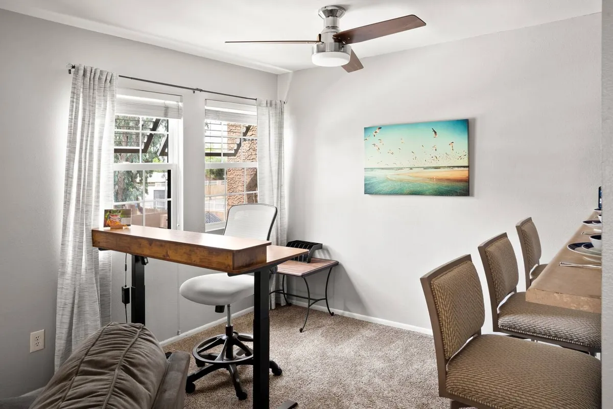 The image showcases a bright, modern condo interior with a sleek wooden bar table and matching bar stools, inviting wall art, and large windows with curtains that let in abundant natural light. The atmosphere feels both productive and relaxing, suggesting opportunities for working, dining, and socializing in a stylish, sunlit space.