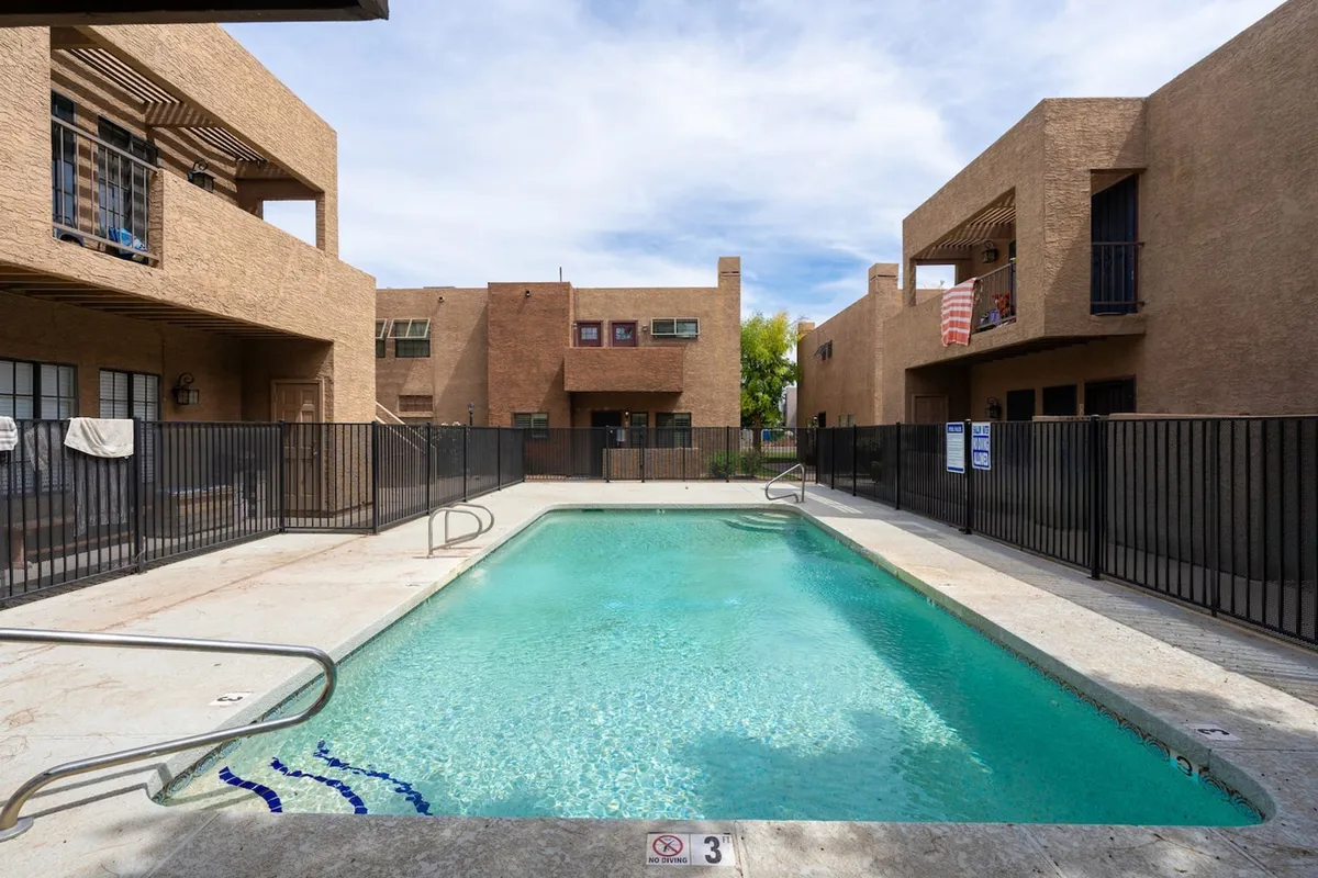 A sunlit exterior view of a resort-style swimming pool surrounded by modern buildings and patio areas. The pool area features ample space for sunbathing and relaxing, with inviting blue water and lounge seating.